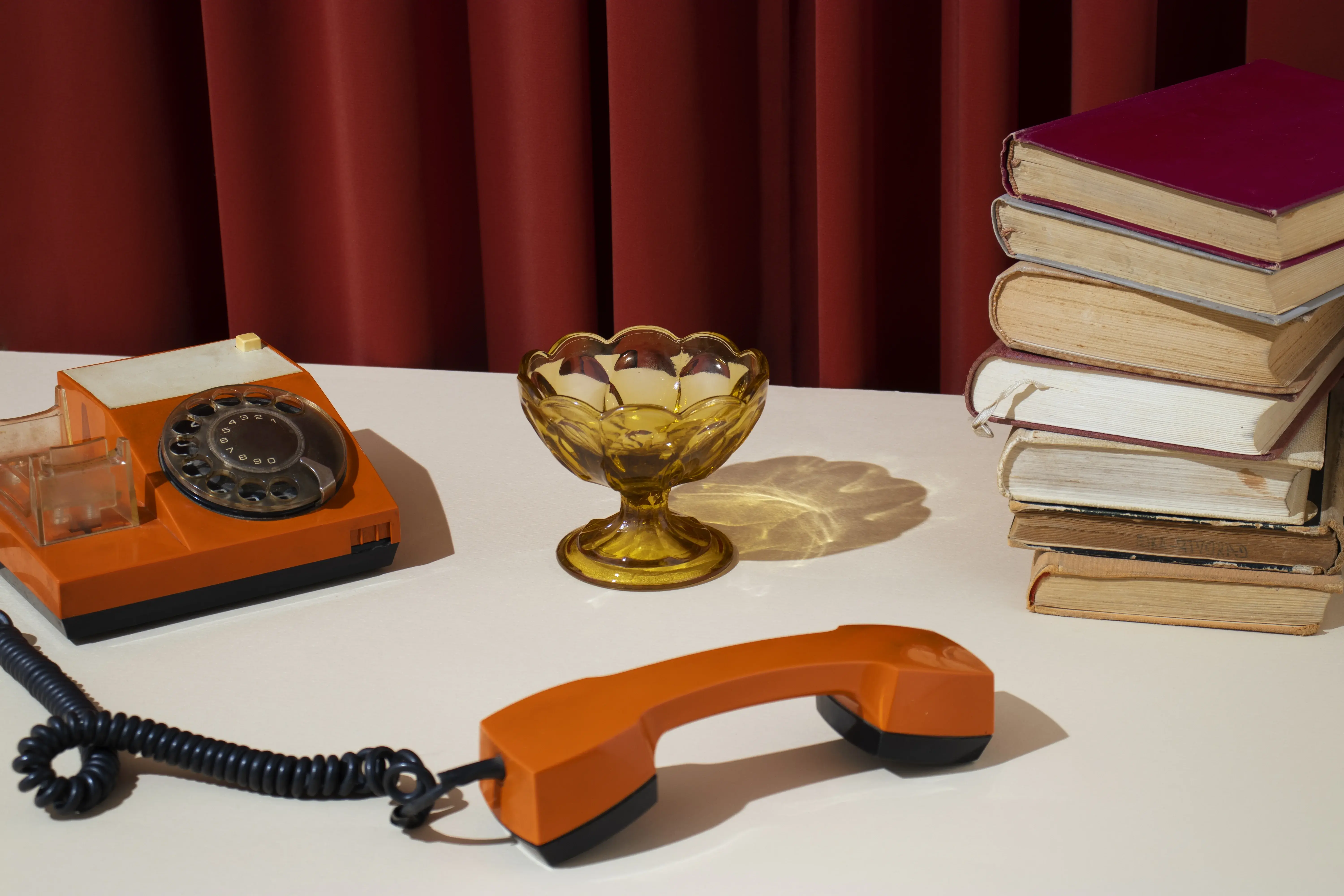 Pomarańczowy telefon stacjonarny, szklany pucharek i stos książek na białym stole.